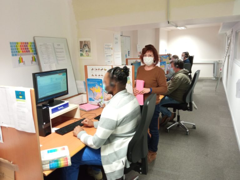 Plusieurs persones sont assis chacun devant un écran dans un centre d'appel avec un casque audio sur la tête. Une femme est debout tenant des fiches roses