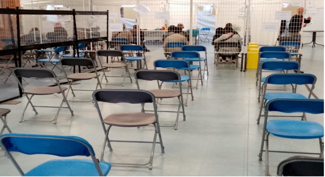 Photo dans un centre de vaccination, des rangées de chaises vides au premier plan, des personnes assises en train d'attendre en arrière plan